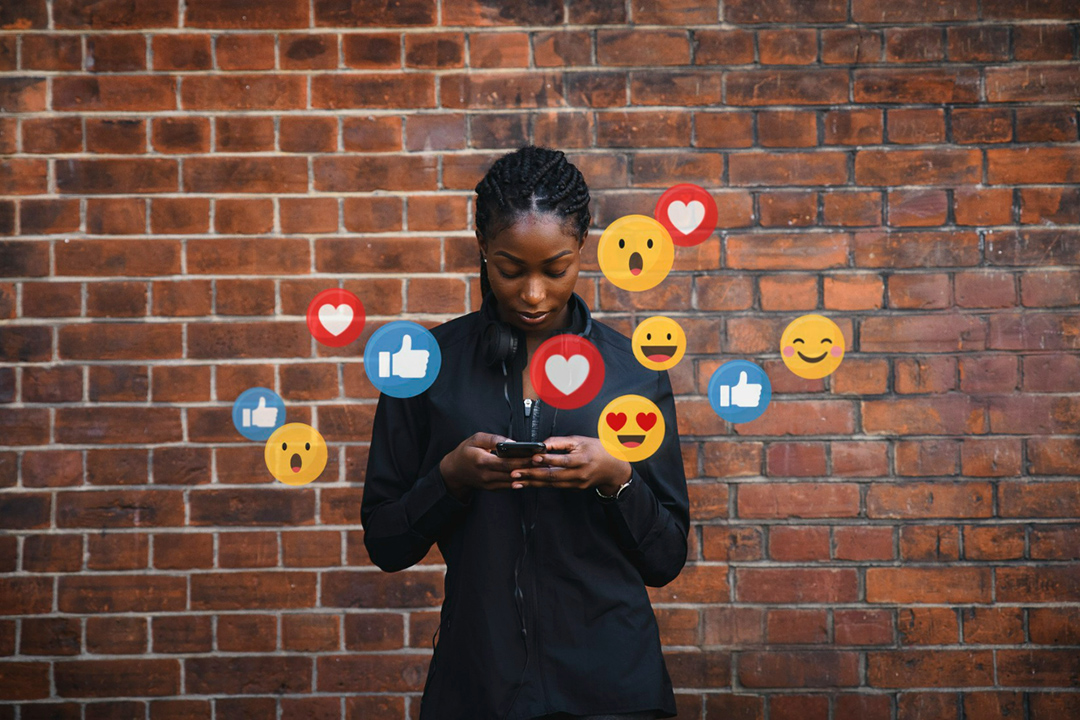 A young woman holding a phone with social media emojis popping out of the screen.